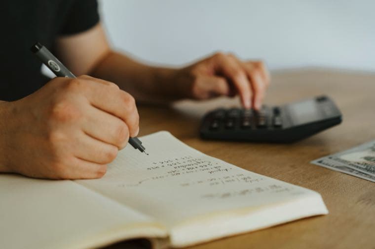 Hands writing budget calculations in a notebook beside a calculator