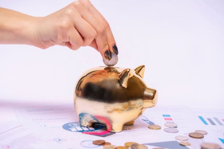 A hand placing a coin into a piggy bank sitting on financial charts